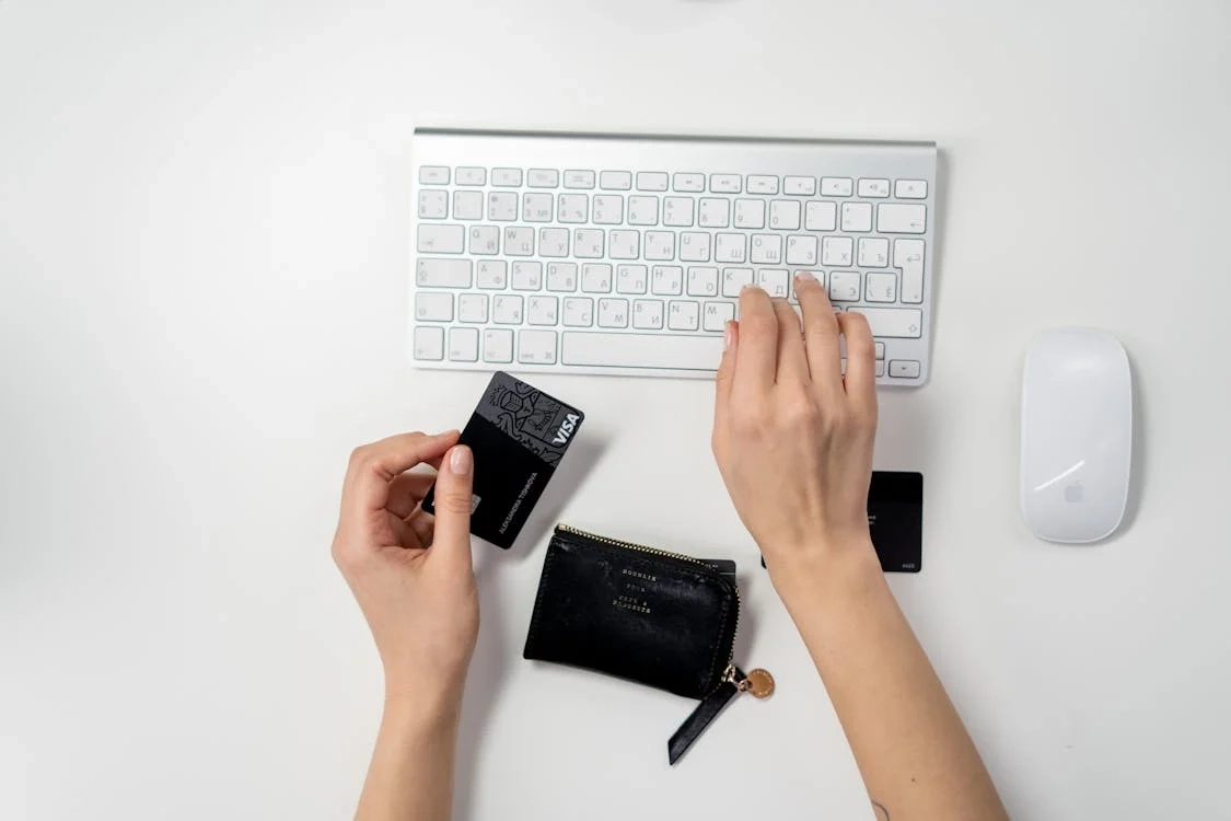 A person making an online payment using a credit card and keyboard on a white desk.
